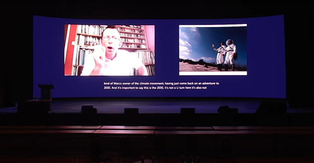 Screenshot of a YouTube video of the talk. On the left, is an image of the speaker mid-sentence. On the right is a still image of the speaker and his colleague dressed in spacesuits pretending to be time travelers