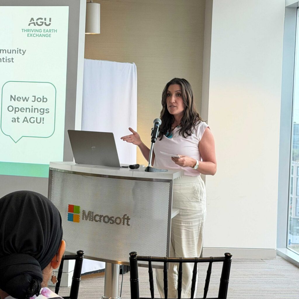 A woman stands in front of a podium and screen with her hands outstretched as she speaks