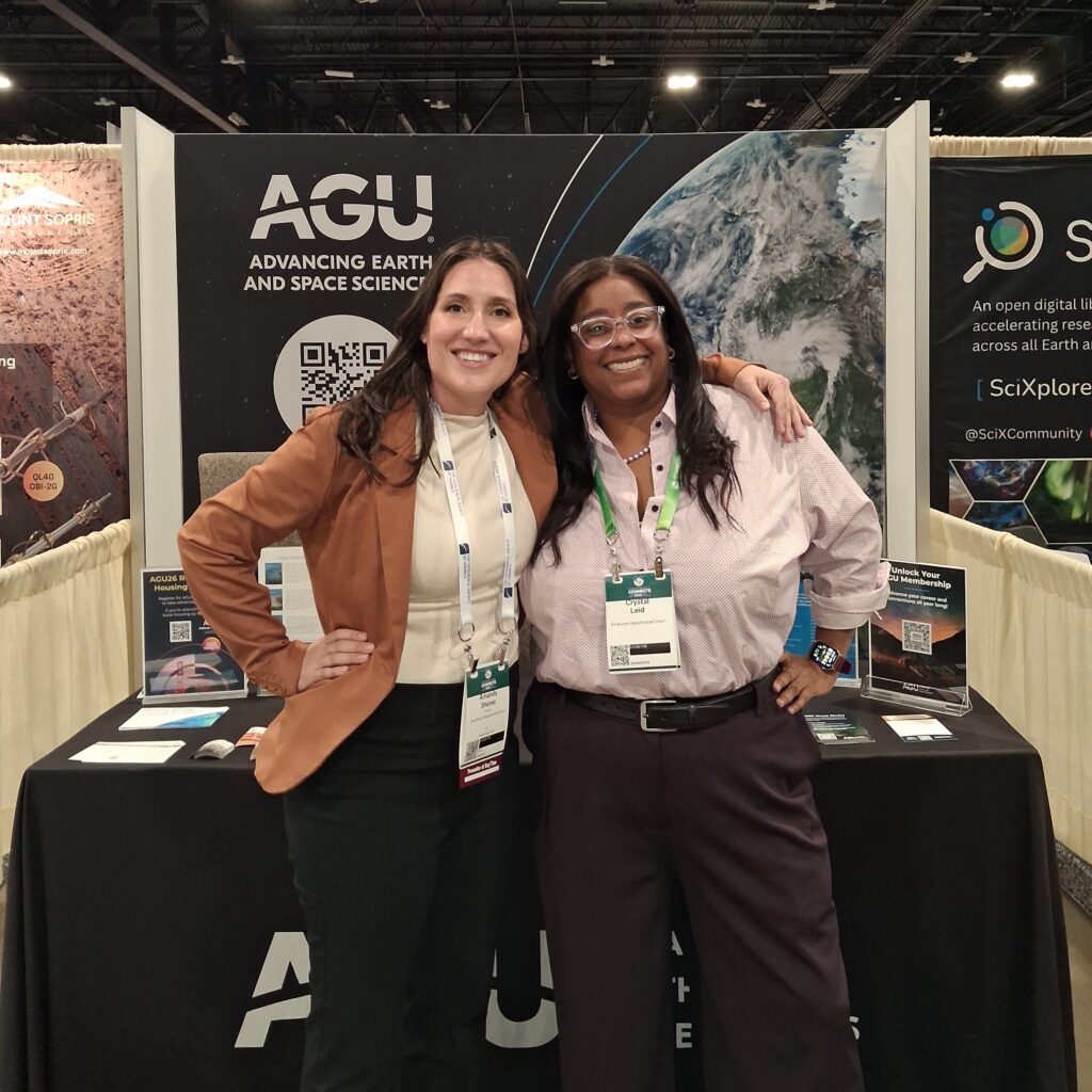 Two women stand smiling in front of the AGU exhibit hall booth