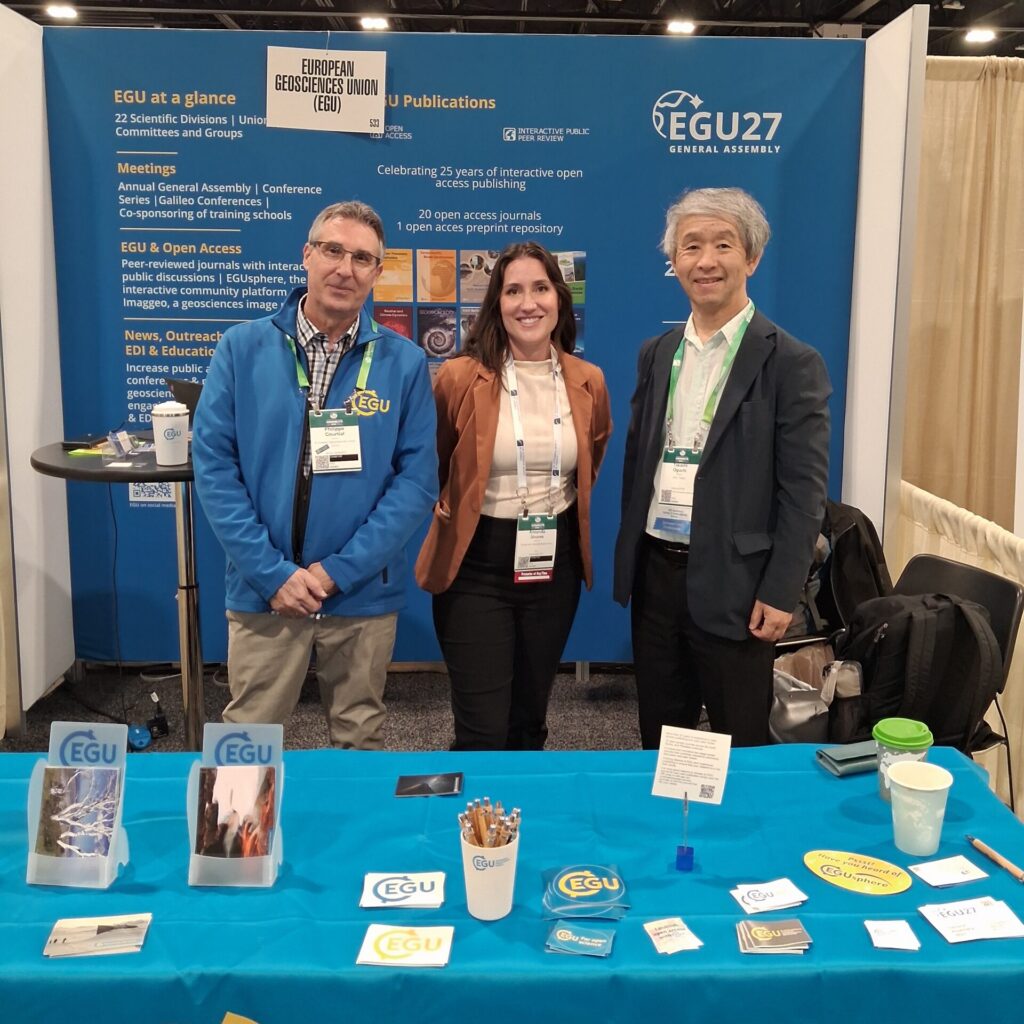 A woman with a white shirt and brown jacket stands between two men at a EGU exhibit hall booth